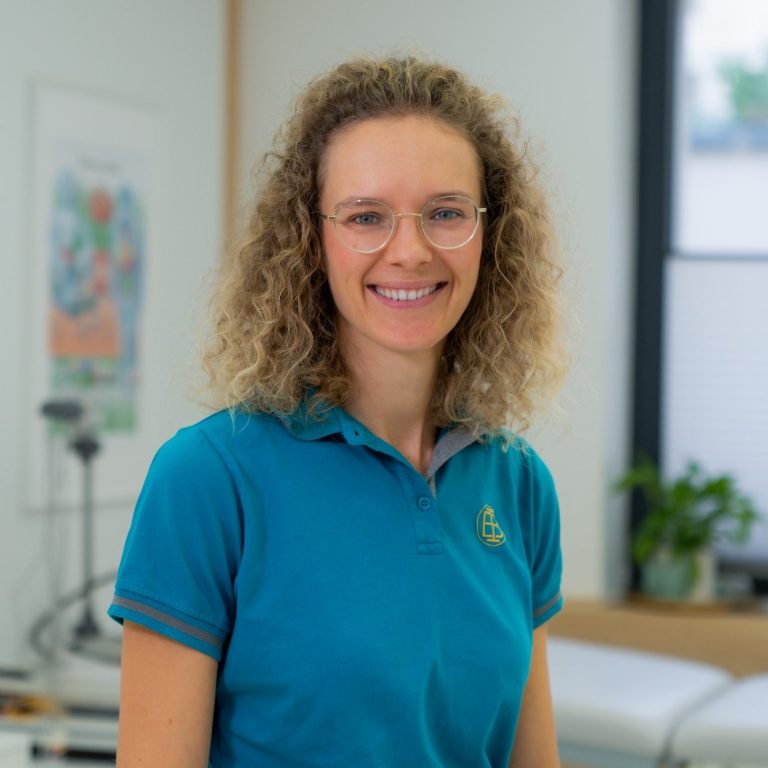 Dr.Anke Müller Frau mit lockigem Haar in blauer Bluse, im Hintergrund Pflanzen und ein Fenster.