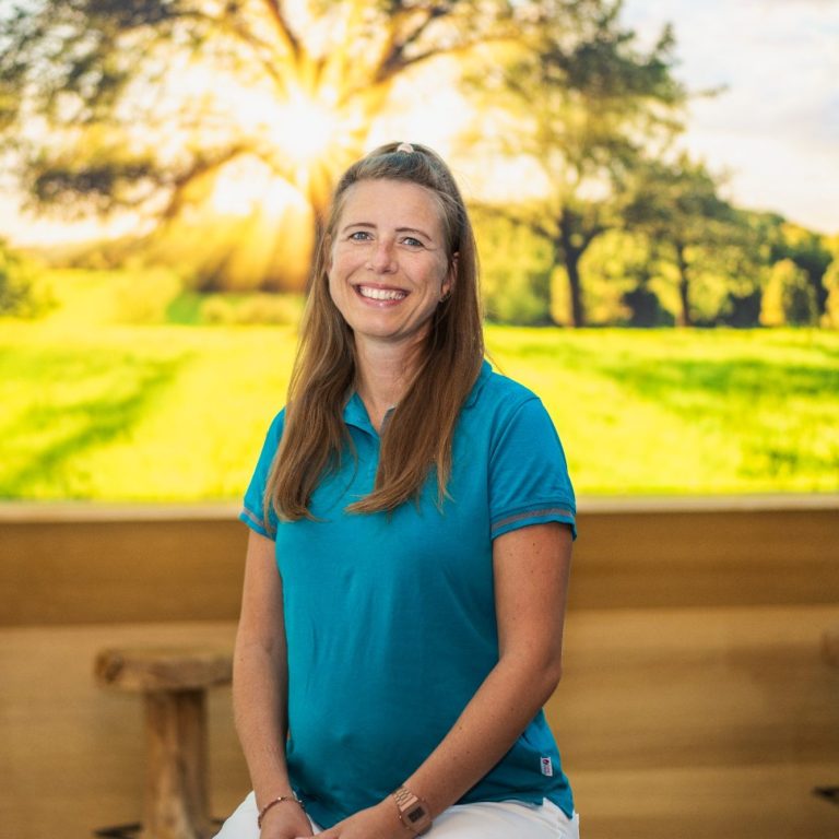 Anne Tusche mit langen Haaren in blauer Bluse sitzt vor einem sonnigen Landschaftshintergrund.