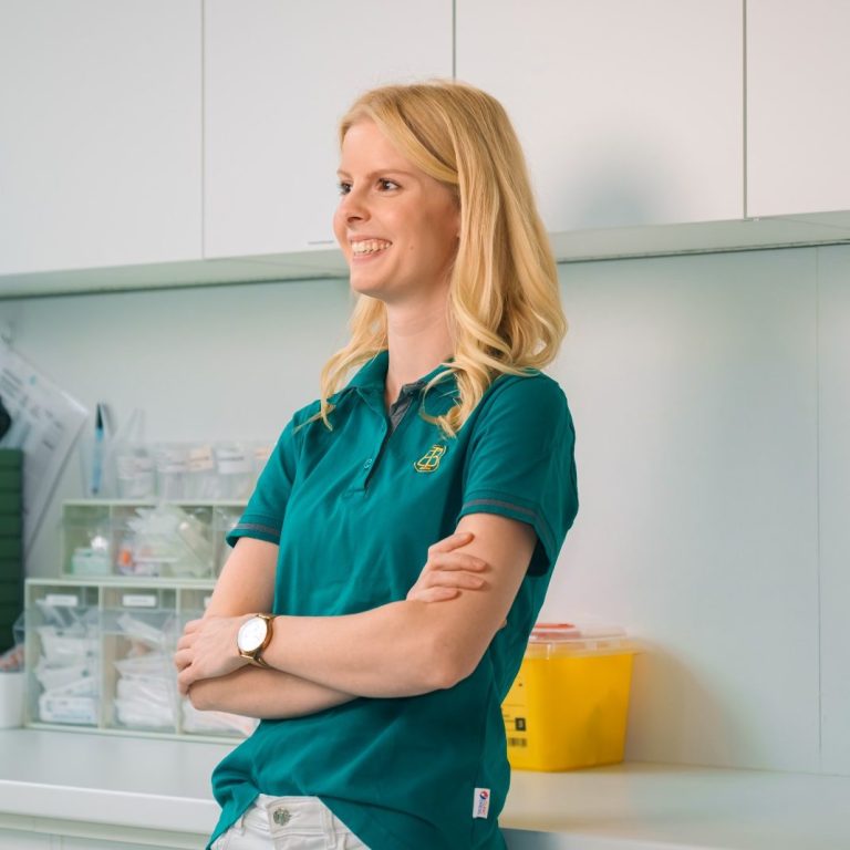 Frau mit blonden Haaren in grünem Polohemd, lächelnd vor einer modernen Arbeitsumgebung. 